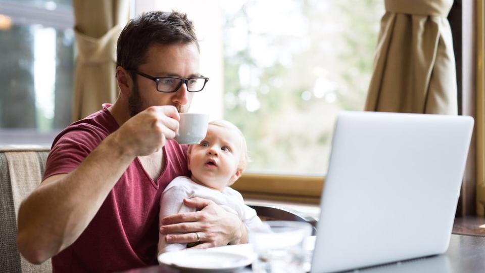 Hombre tomando un café, con un bebé en brazos frente a una computadora.