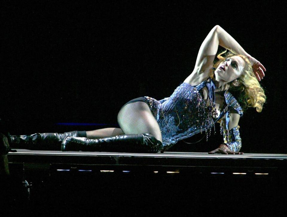 madonna, wearing a blue outfit, fishnet stockings, and black leather boots, lies on a stage while looking up, with her right arm resting on her head