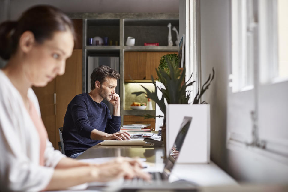 Mid adult man using computer. Woman is working in foreground at desk. Business couple are in home office.