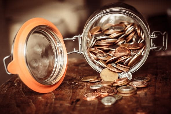 A jar of sterling coins is tipped on its side with the coins spilling out on to a wooden surface. The jar denotes a savings pot.