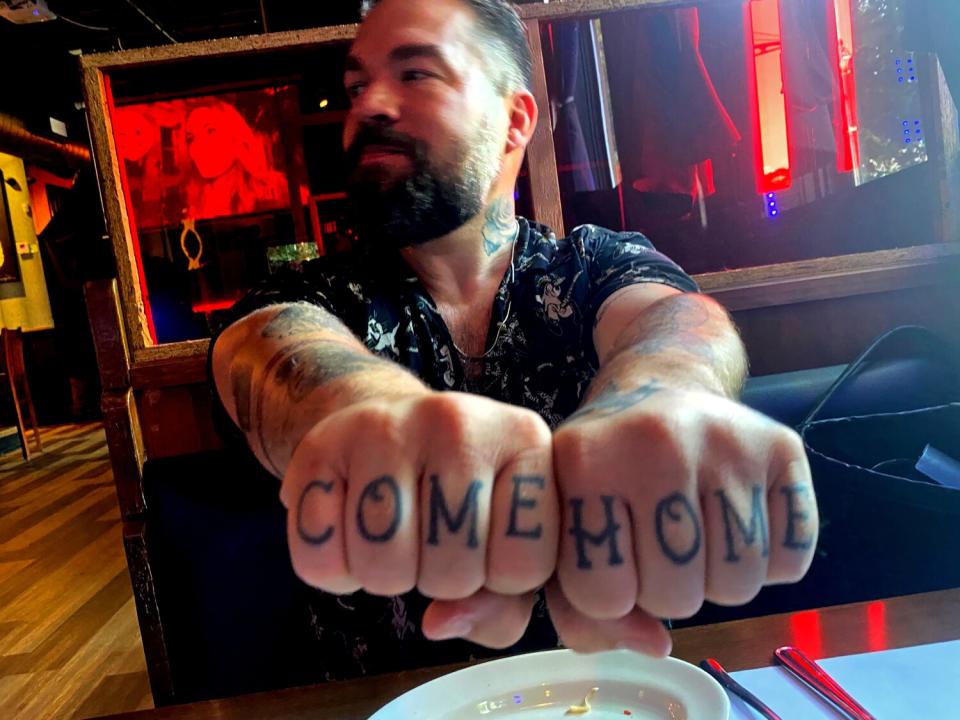 A man seated at a table holds out his hands to display the words "come home" tattooed on his knuckles.