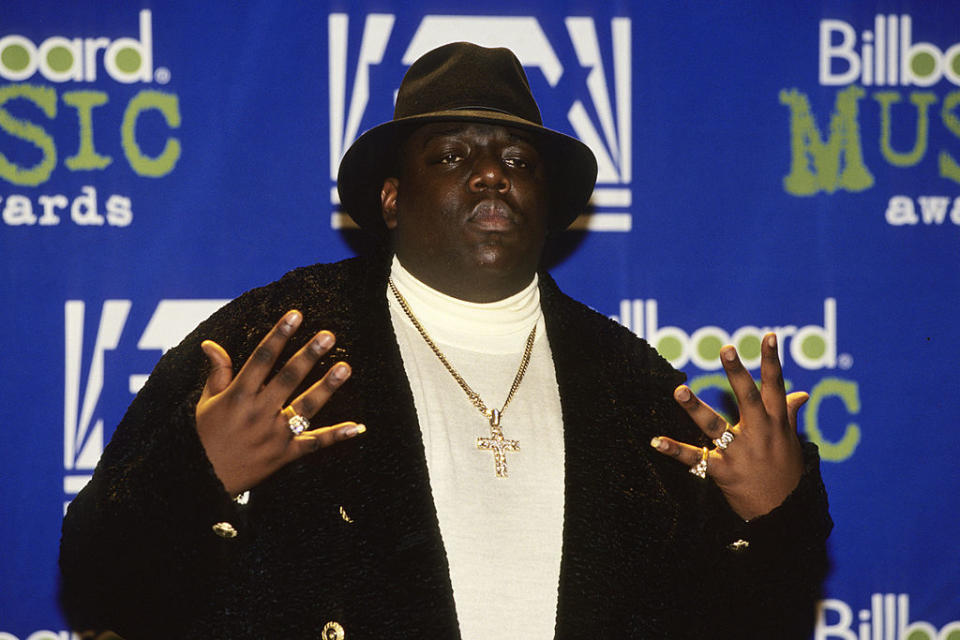 The Notorious B.I.G. poses with hands raised, wearing a fur coat, fedora, white turtleneck, gold chains, and rings at the Billboard Music Awards