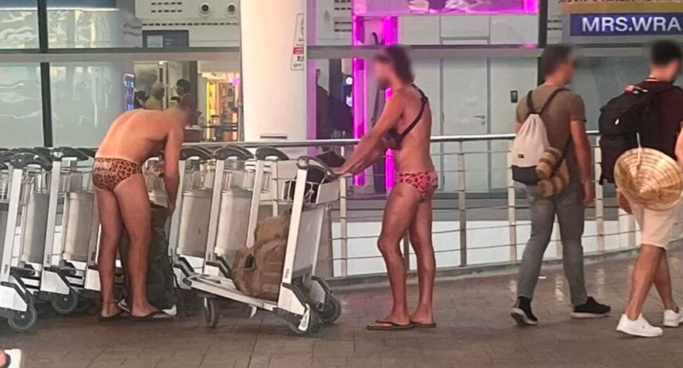 Two men in Speedos at Phuket International Airport putting luggage on a trolley. 