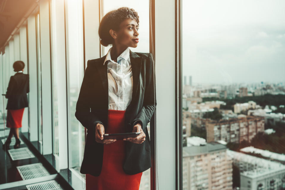 An African-American woman entrepreneur a digital tablet is leaning against a panoramic window of an office business skyscraper and looking in a distance; biracial businesswoman with a tablet pc