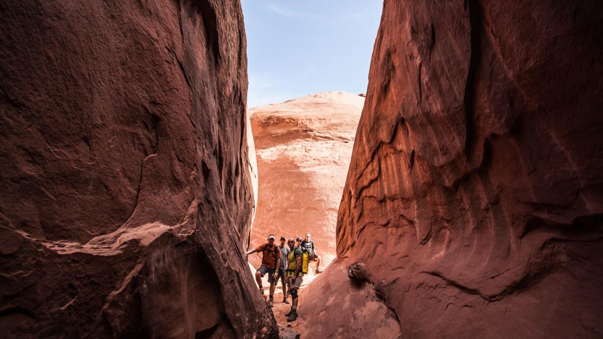 five men stand at the base of a slot canyon with sandstone walls and a clear blue sky beyond.