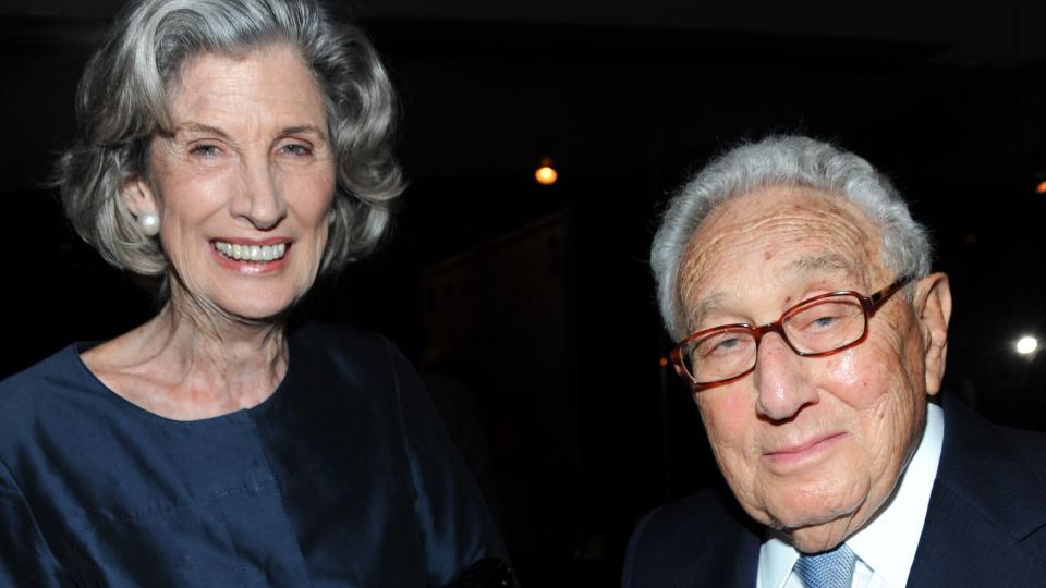 nancy kissinger and henry kissinger stand next to each other and smile for a photo, she wears a blue long sleeve gown, he wears a navy suit jacket, light blue tie, and white collared shirt