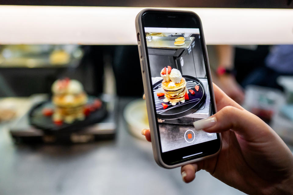 A person holds a phone, capturing a photo of a plate of pancakes with whipped cream and strawberries