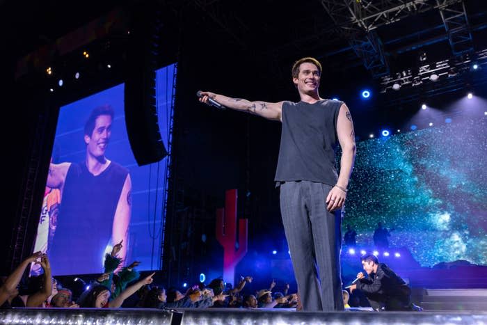 Nicholas Galitzine on stage in The Idea of You smiling with a microphone, background screen shows their image, crowd in foreground