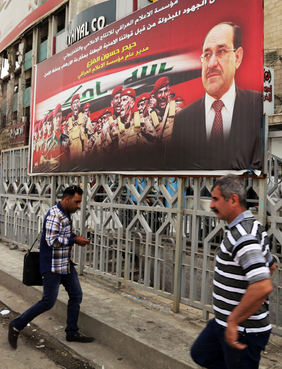 In this Tuesday, March 18, 2014 photo, Iraqis walk past an anti-terrorism banner with a photo of Iraqi Prime Minister Nouri al-Maliki, in Baghdad, Iraq. Islamic militants who took over the Iraqi city of Fallujah are now trying to show they can run it, providing social services, policing the streets and implementing Shariah rulings in a bid to win the support of its Sunni Muslim population. Partial translation of Arabic on banner reads, "the media supports security forces efforts to capture terrorists and resume safety." (AP Photo/Karim Kadim)