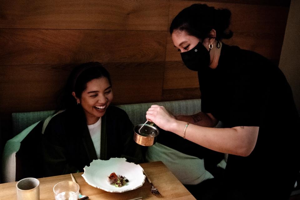A woman prepares a dish table side for another woman that is smiling.