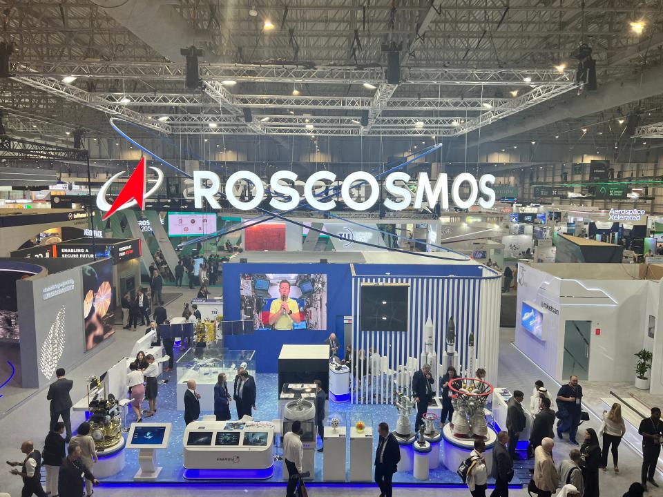 The Roscosmos stand at the Dubai Airshow viewed from high-up, has a large neon sign suspended from the ceiling while people look at satellites on display