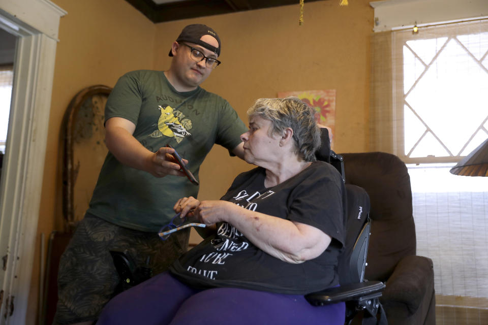 In this Tuesday, April 28, 2020, photo Zach Stafford, left, shows his mother, Debra Mize, a news alert on his phone as they watch a livestream of the daily coronavirus briefing by Illinois Gov. J.B. Pritzker on a television just out of view inside their home in Belleville, Ill. The pair say they are consuming hours of news each day in various formats about the coronavirus. Americans are grappling with an essential question as they try to get the information they need to stay safe during the coronavirus crisis: Whom do you trust? (AP Photo/Jeff Roberson)