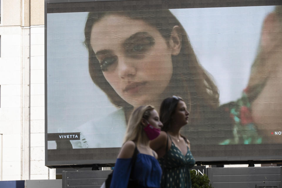 Pedestrians pass by a screen showing a Vivetta model during the Milan Digital Fashion Week, in Milan, Italy, Tuesday, July 14, 2020. Forty fashion houses are presenting previews of menswear looks for next spring and summer and pre-collections for women in digital formats, due to concerns generated by the COVID-19. (AP Photo/Luca Bruno)