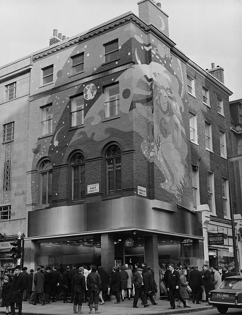 A black-and-white photograph of a busy street scene featuring a building with a large mural of a cosmic figure on the facade