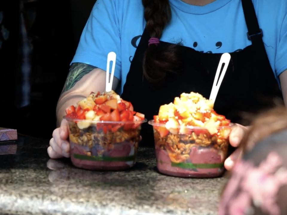 A worker is holding two açaí smoothie bowls, which are topped with a variety of fruits and nuts.