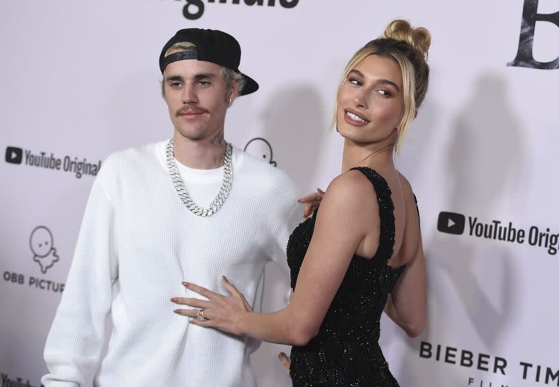 Justin Bieber, dressed in white long sleeve and Hailey Bieber in black dress smiling together on red carpet