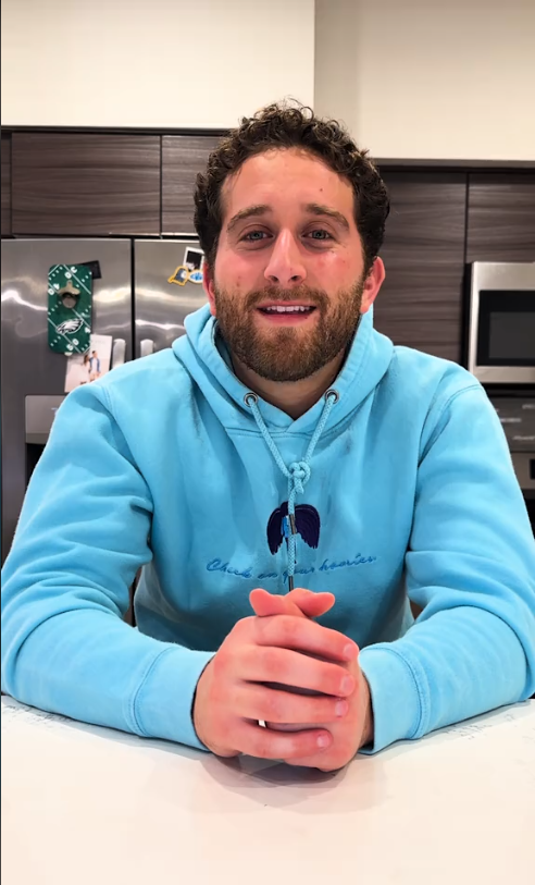 A man in a casual hoodie sits at a kitchen counter, smiling