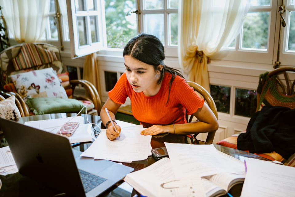 hispanic latina college student works on assignment in her dorm room. She is writing something in a notebook. A laptop is on her desk.