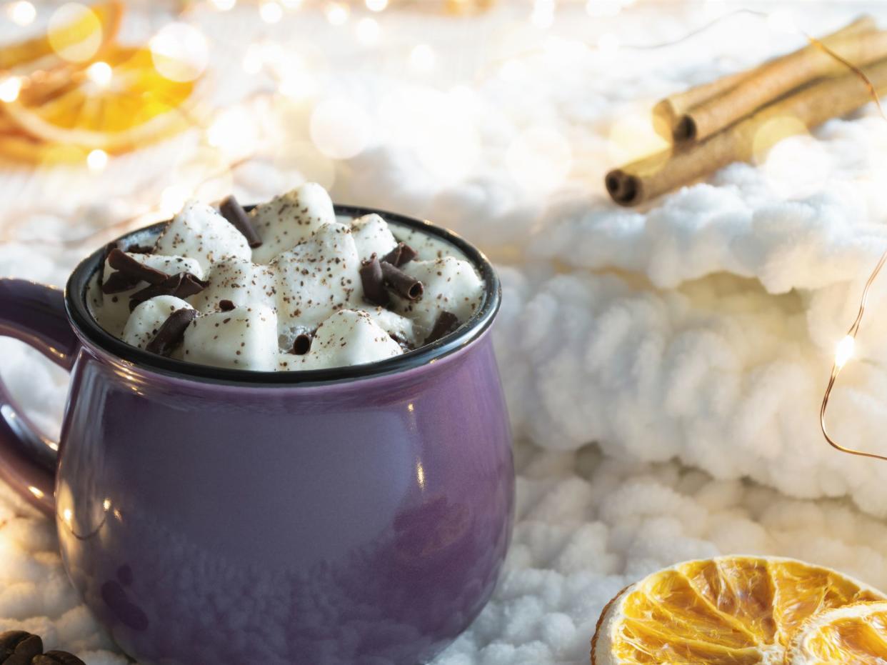 Hot cocoa with marshmallows, slices of dried orange and cinnamon sticks on knitted fabric. Christmas lights in the background. Cozy holidays concept.