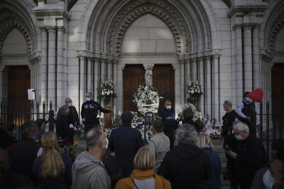 People gather as flowers, messages and candles lie in front of the Notre Dame church, in Nice, France, Friday, Oct. 30, 2020. A new suspect is in custody in the investigation into a gruesome attack by a Tunisian man who killed three people in a French church. France heightened its security alert amid religious and geopolitical tensions around cartoons mocking the Muslim prophet. (AP Photo/Daniel Cole)