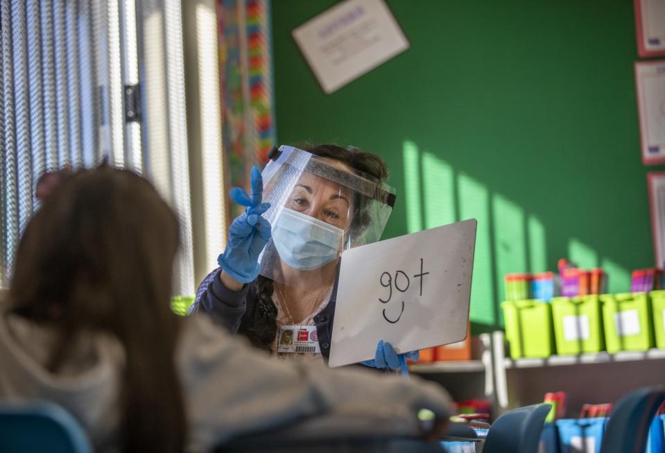 A teacher's aide wearing a face mask and shield gives a lesson