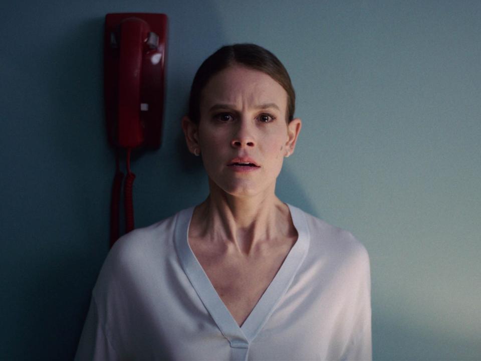 Sosie Bacon standing in front of a wall with a red telephone handing on her right side a distraught expression on her face in the movie "Smile."