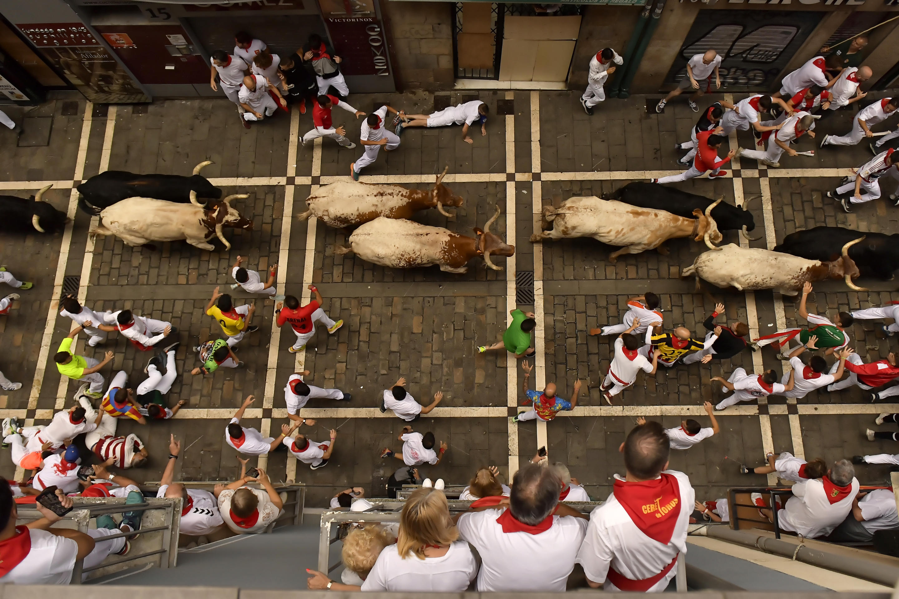 A view from above of bulls and festivalgoers running down a narrow street during the Pamplona bull run as spectators watch from a balcony.