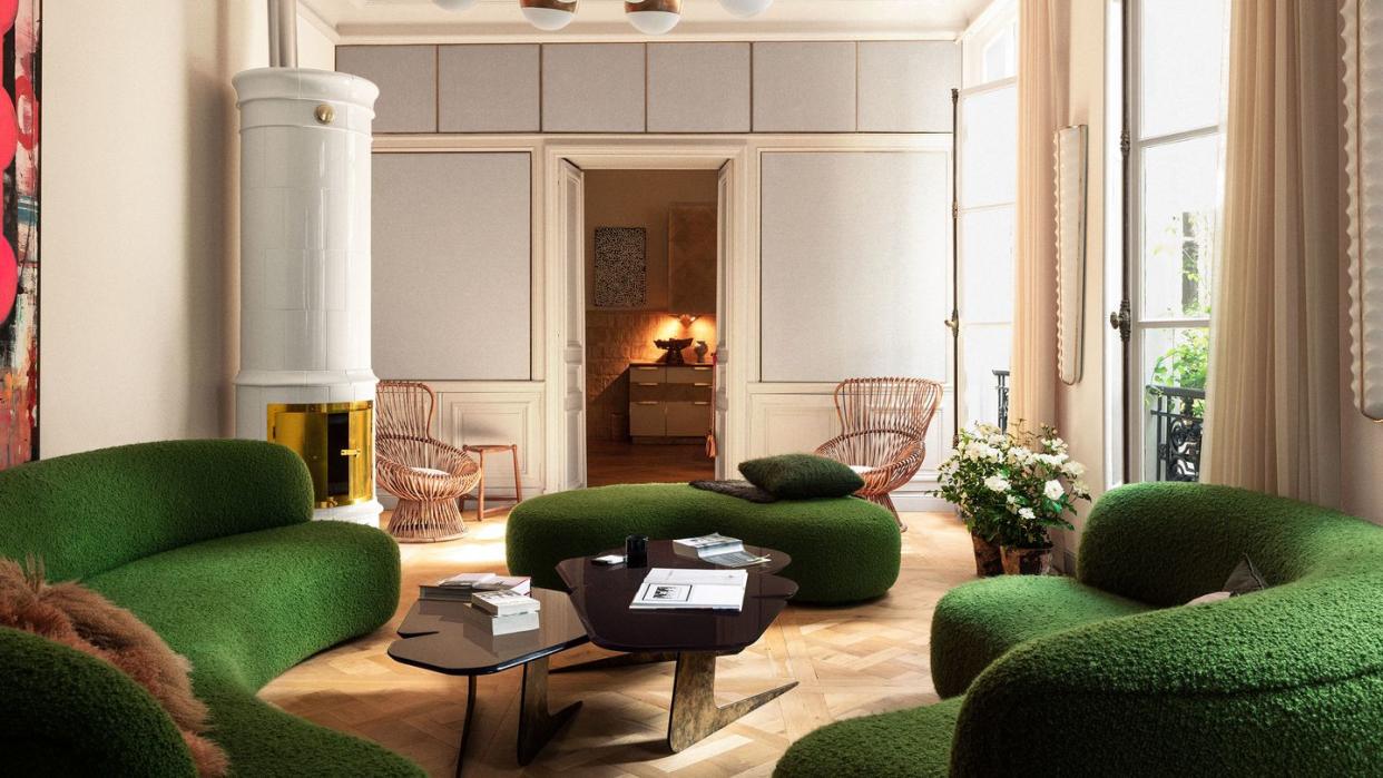 expansive european looking living space with multiple curvy seating sofas in a lush dark green and an extravagant but simple chandelier of multiple white builbs with gold bands and a double tier cocktail table