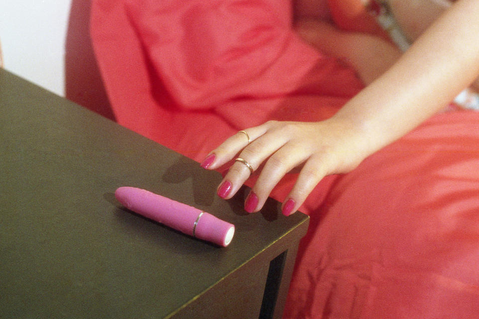 A person's hand with rings resting next to a pink object on a dark surface, against a red fabric background