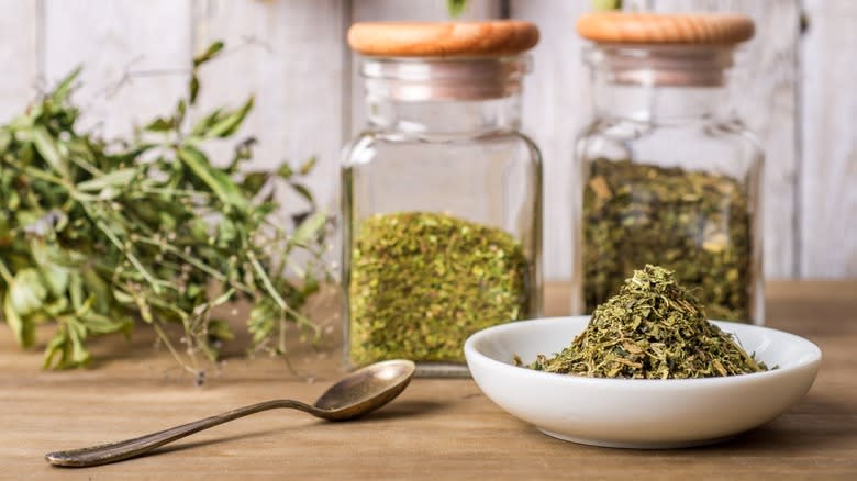 Close-up of dried herbs in a bowl next to a spoon and glass jars of herbs