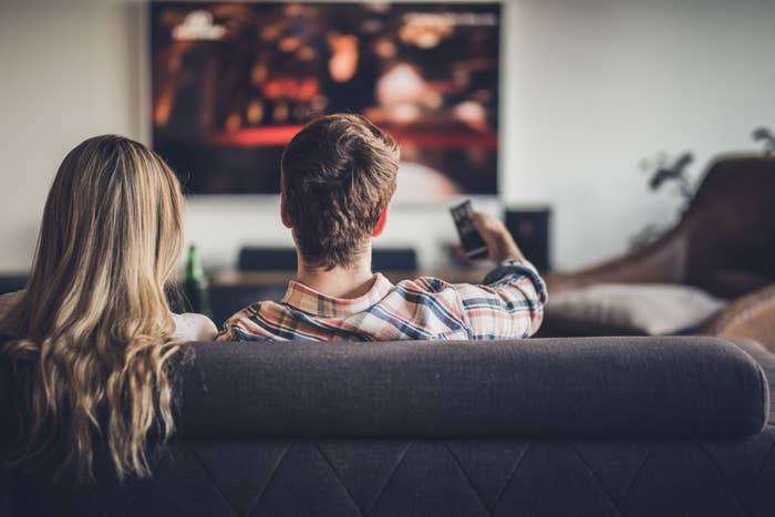 A man and a woman are sitting on a couch facing a television, with the man holding a remote control