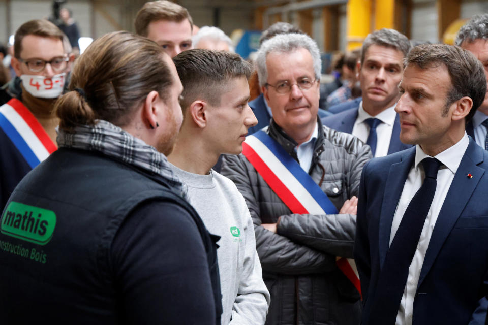 French Parliament member Emmanuel Fernandes, left, of the France's Unbowed party, wear a gag over his mouth showing his opposition to the pension reform, appears as French President Emmanuel Macron, right, talks to employees a beer during a visit to Mathis, a company specialized in large wooden buildings, in Muttersholtz, eastern France, Wednesday, April 19, 2023. (Ludovic Marin, Pool via AP)