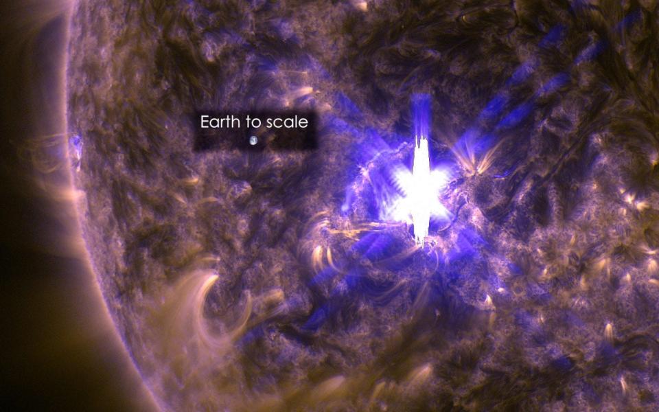 the sun up close in purple ultraviolet light with a bright purple and white star shaped solar flare erupting from the surface and earth superimposed nearby for scale much smaller than the solar flare