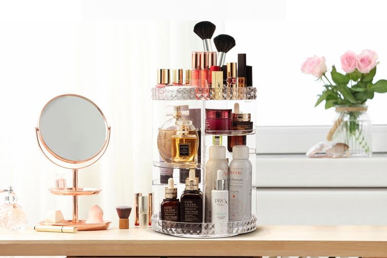 A dressing table with makeup and cosmetics arranged on an Estelle Diamante 360° Rotating Adjustable Makeup Organiser with a small oval mirror and pink roses in a vase.