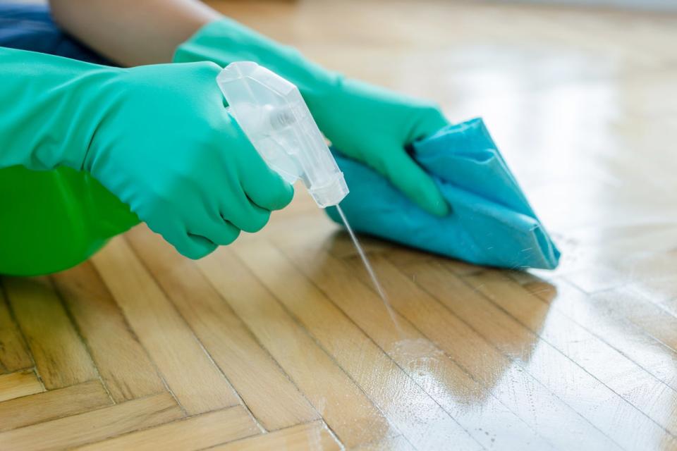 A person in green gloves sprays a cleaning solution on a hardwood floor.