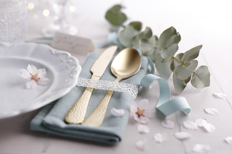 Gold-plated cutlery wrapped in lace ribbon on a blue napkin, with a white plate and floral decorations, suitable for a wedding table setting