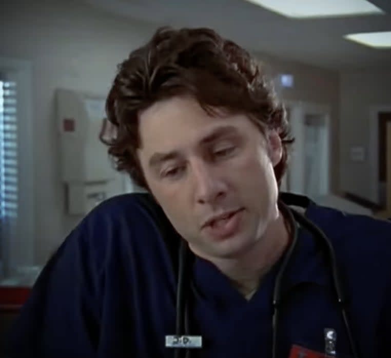 Zach Braff in a hospital setting wearing a dark scrub top with an ID badge and a stethoscope around his neck