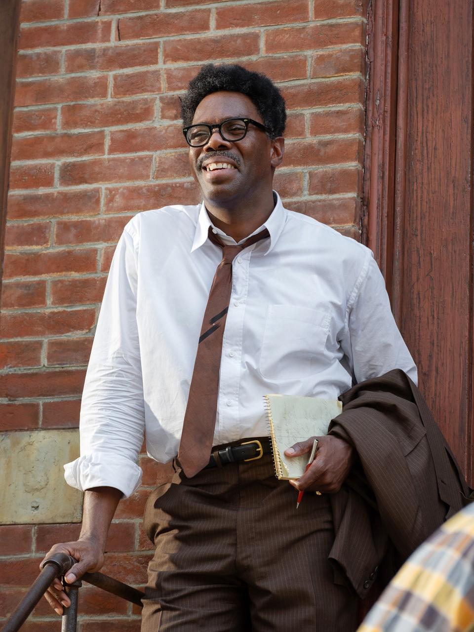 Colman Domingo as Bayard Rustin smiling and leaning onto a stair handrail outside, wearing a dress shirt, loose tie, and slacks