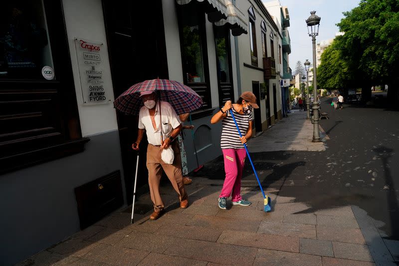 A man walks with an umbrella as women clean the ash from the street following the eruption of a volcano on the Canary Island of La Palma