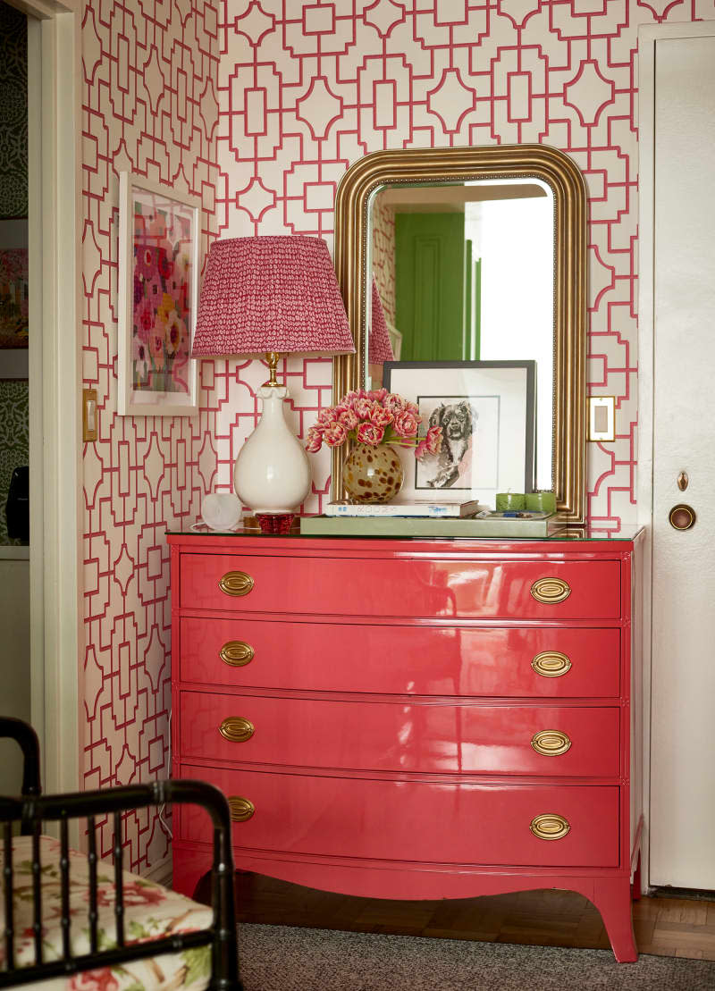 Mirror, art, flowers, and lamp atop pink dresser in wallpapered entryway.