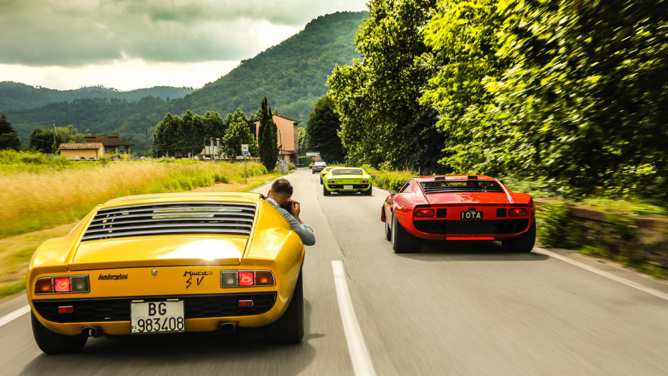 Italian supercars roll down a rural road.