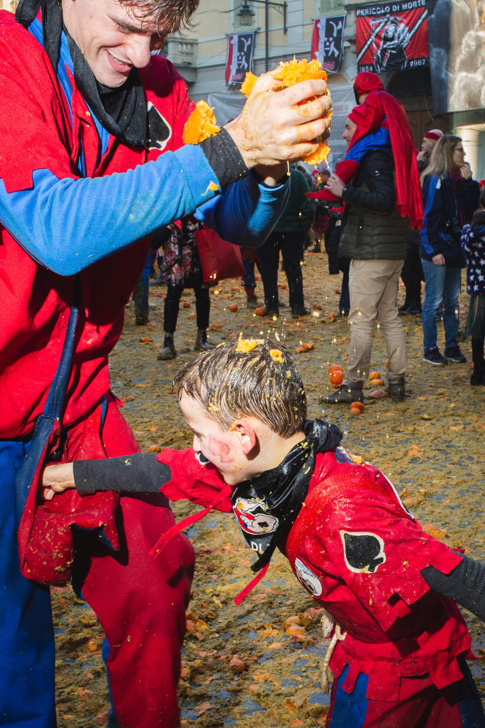 A man holds two big fistfuls of crushed oranges, spilling onto a young boy punching the man's groin