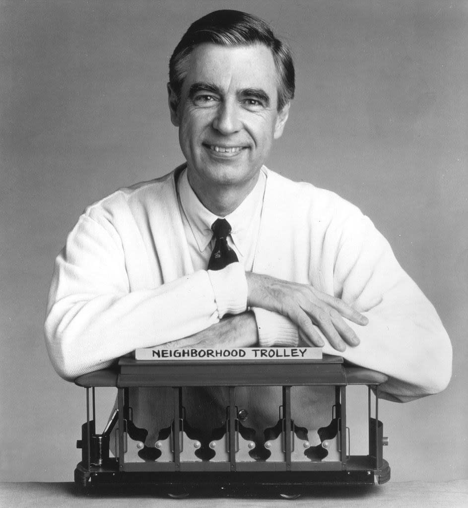 Mr. Rogers smiling with his hands folded across a model train from his show