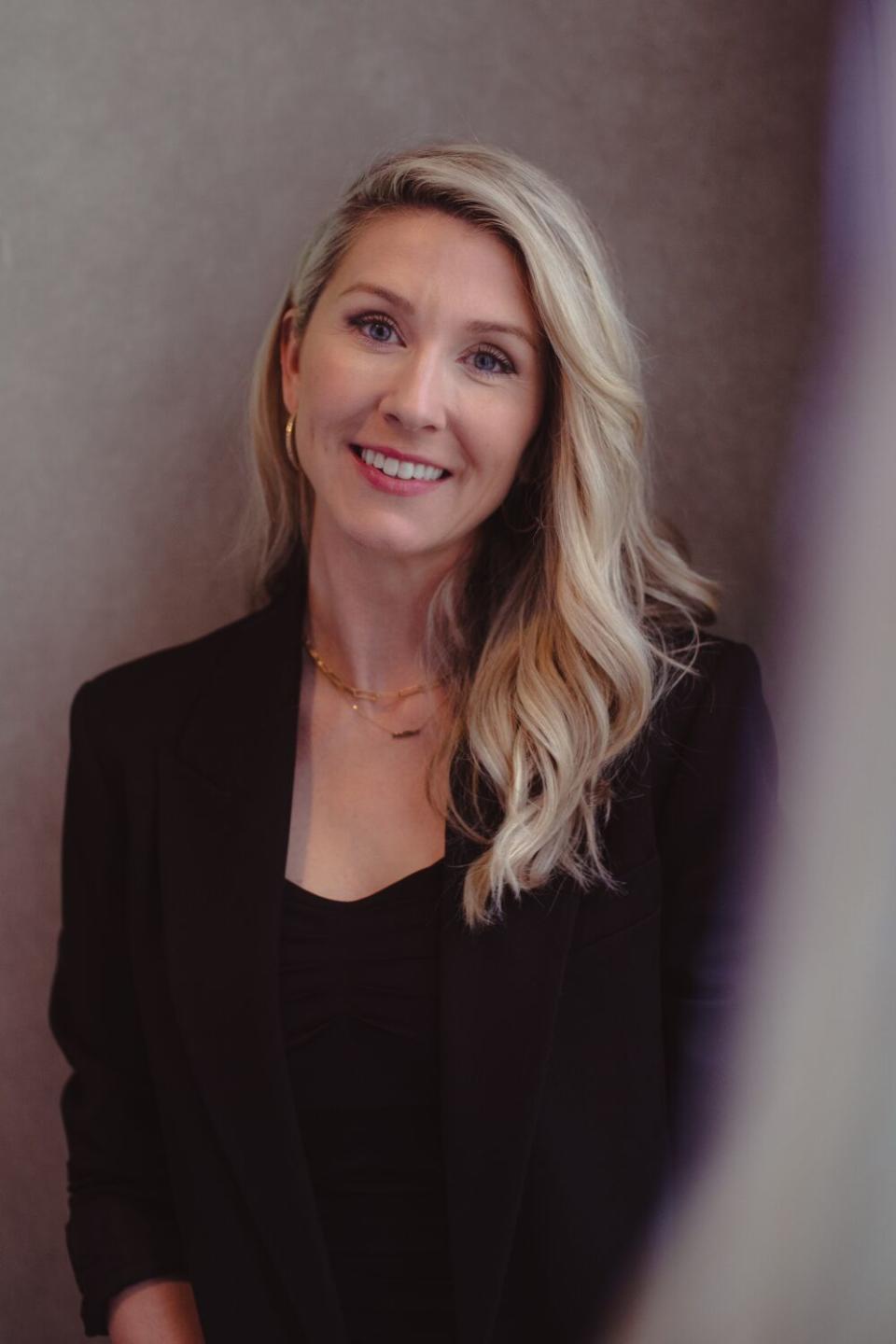 A blonde woman in a black shirt and blazer against a grey backdrop.