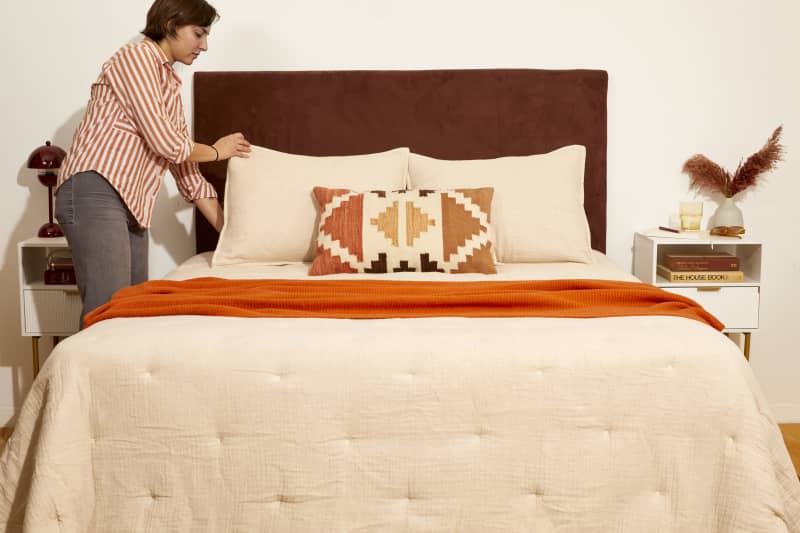 Head on shot of a woman in a red and white striped shirt, adjusting the beige pillows on a made bed with the new DIY headboard hanging behind it.