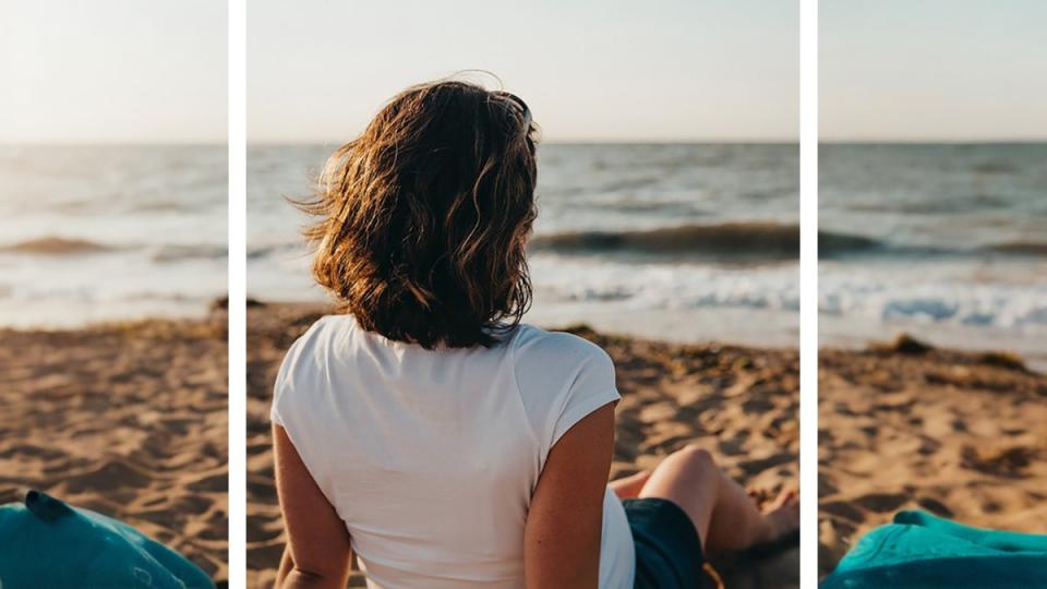 A woman with shoulder-length brown hair sitting on a beach in a white t-shirt and black shorts, with additional landscape and some teal beach towels to the left and right created by Clipdrop's Uncrop, an AI image extender tool.