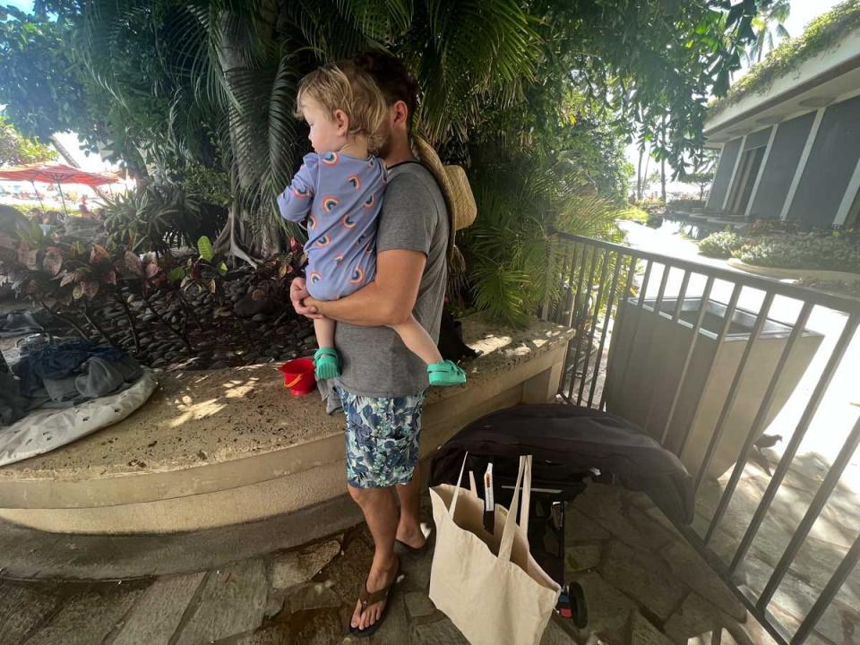 A man standing holding a baby next to a tree and a gate and a stroller.