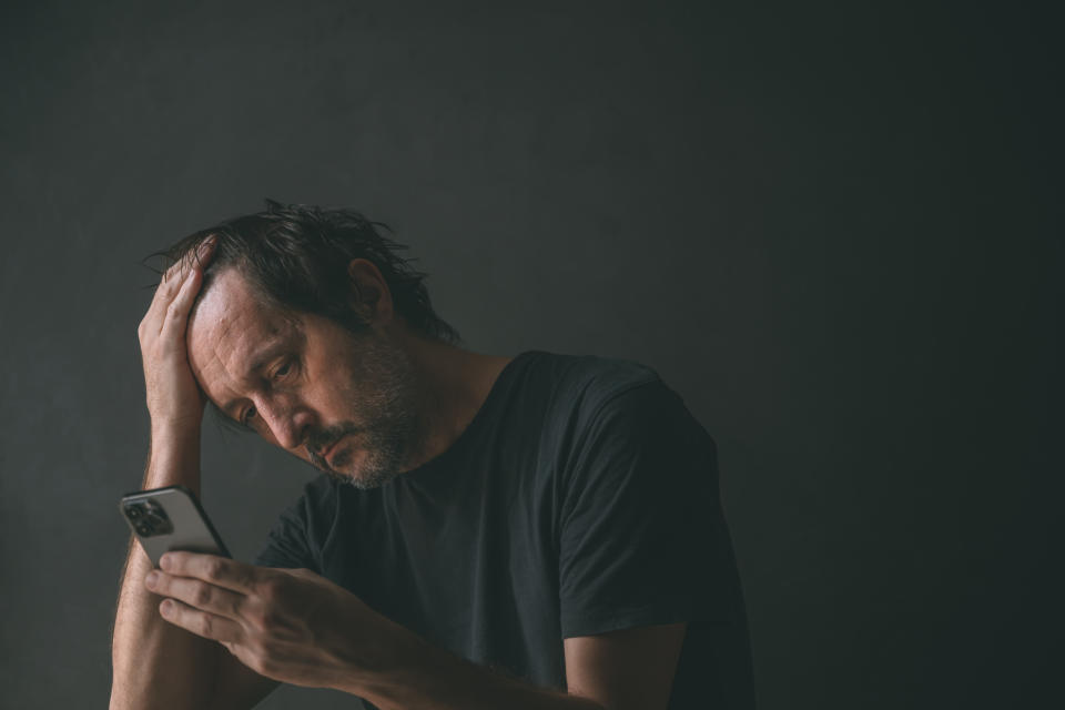 Man in a black t-shirt holds his phone, looking stressed as he runs his other hand through his hair against a dark background