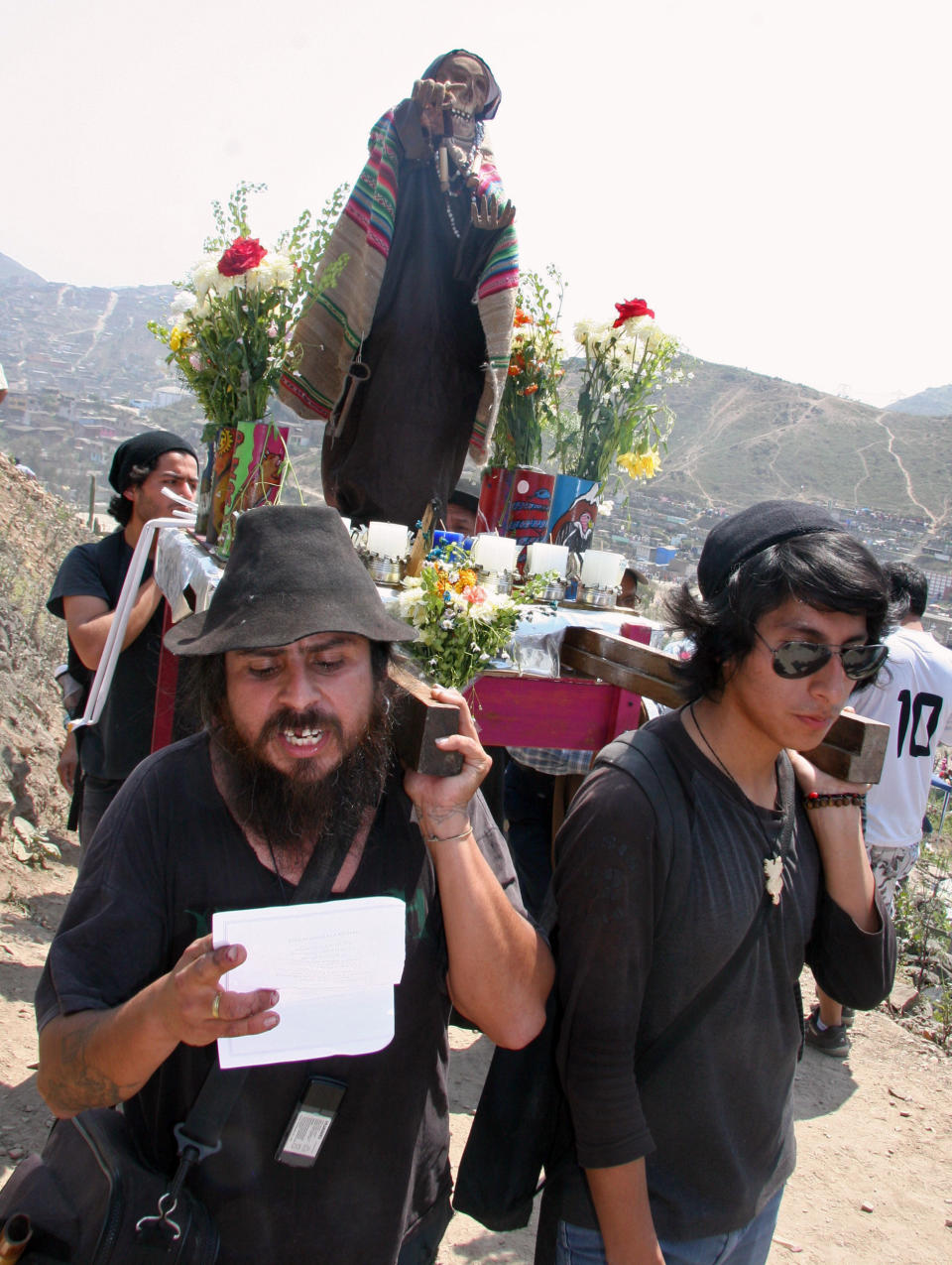 In this Nov. 1, 2012 photo, Fernando Naveda, left, reads a poem dedicated to Santa Muerte, as he and Claudio Ajayu, right, carry a sculpture of the folk saint during the Day of the Dead festival at the Cemetery of Nueva Esperanza in Villa Maria del Triunfo in Lima, Peru. (AP Photo/Jody Kurash)