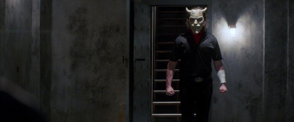 A man with a devil mask stands in a dark room by a staircase